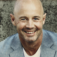 Bald man with a friendly smile, wearing a blue blazer, against a textured gray background.