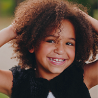 Smiling young child with curly hair, hands playfully on head, showcasing a joyful expression.